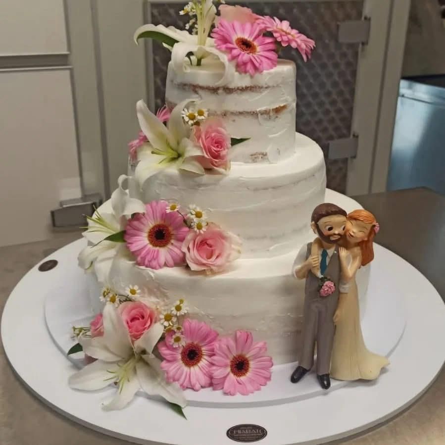 Torta nuziale a tre piani con gerbere rosa, gigli e statuine degli sposi